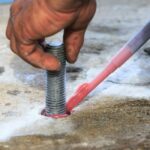 Worker injecting epoxy resin into a drilled hole for chemset anchor bolt installation on a concrete slab.
