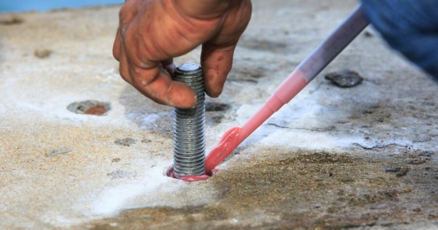 Worker injecting epoxy resin into a drilled hole for chemset anchor bolt installation on a concrete slab.